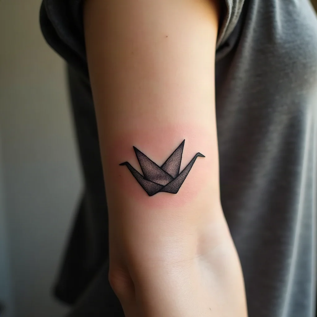 Half a fine-line paper crane tattoo on a mother's inner forearm, designed to complete her son's matching half